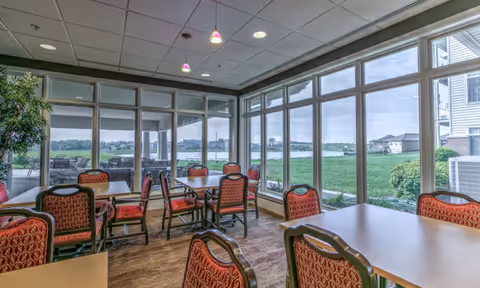 A dining area with multiple tables and red cushioned chairs arranged neatly. Large floor-to-ceiling windows provide a clear view of a green lawn and a body of water outside. The ceiling has recessed lighting and hanging pendant lights.