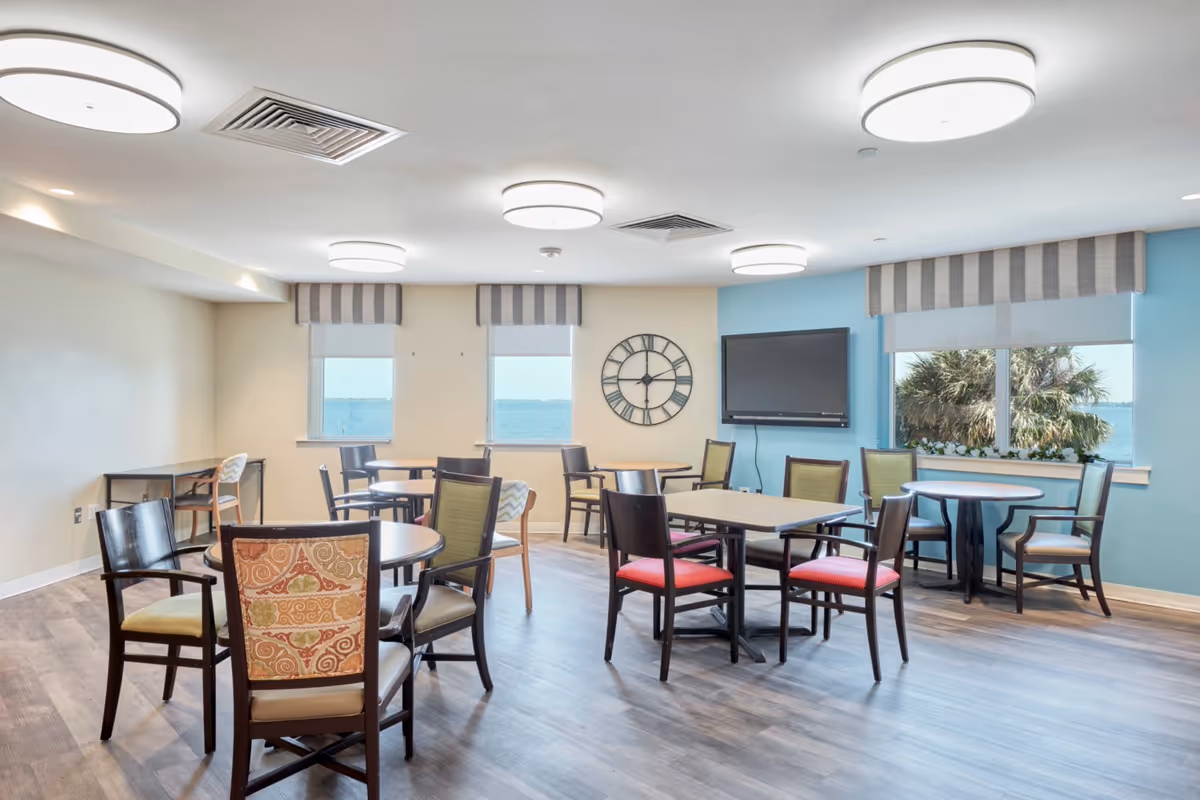 A bright and spacious common area with multiple round and square tables surrounded by chairs. The room has large windows with views of water and palm trees outside. A large wall clock and a flat-screen TV are mounted on the walls. The floor is wood, and the ceiling has several round light fixtures.