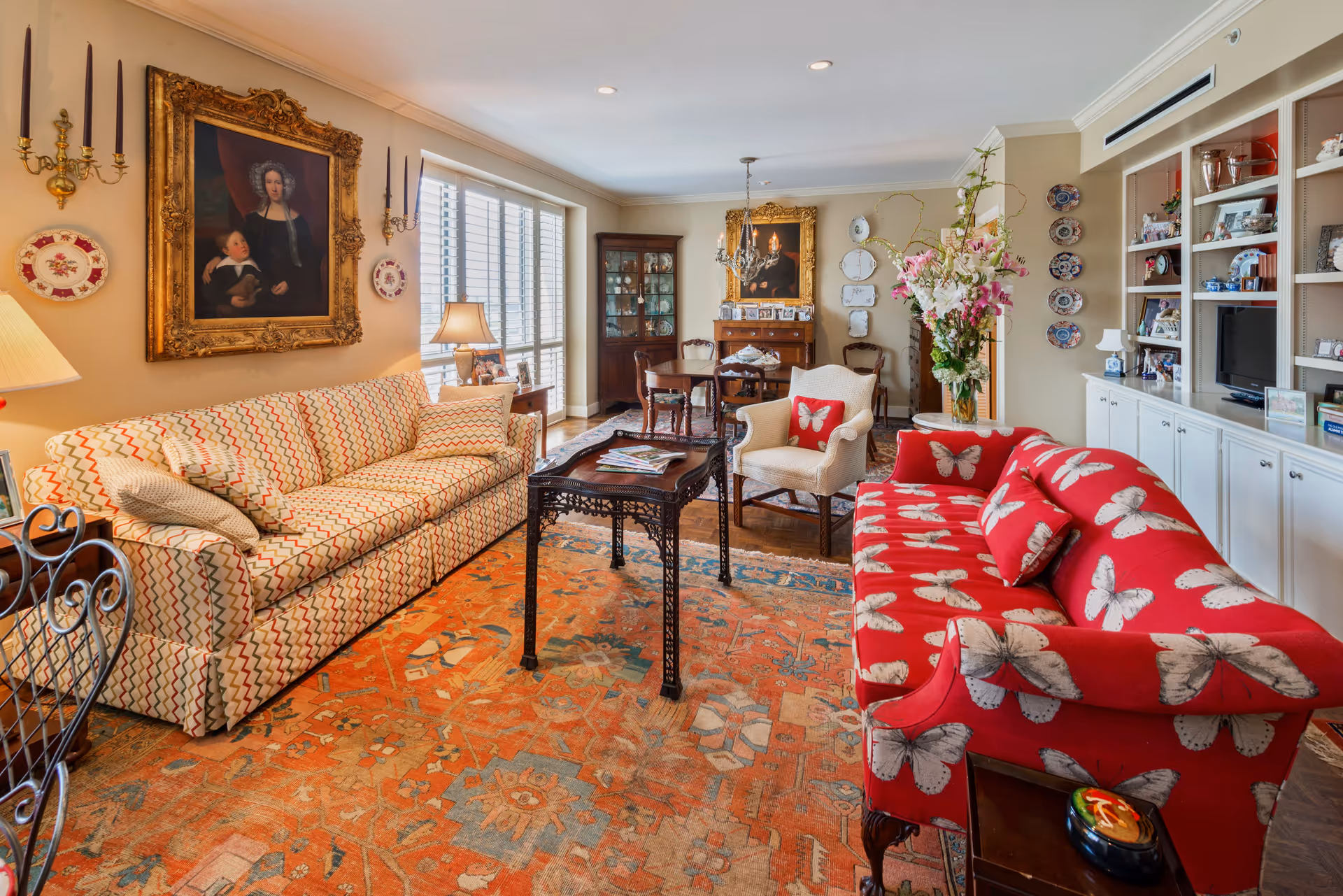 A cozy living room with two patterned sofas, one beige with a zigzag design and the other red with large white butterflies. A wooden coffee table sits on an ornate orange and blue rug. The room features a large window with white shutters, antique-style paintings in ornate gold frames, a chandelier, and built-in white shelves filled with decorative items and books. A dining table with chairs is visible in the background.