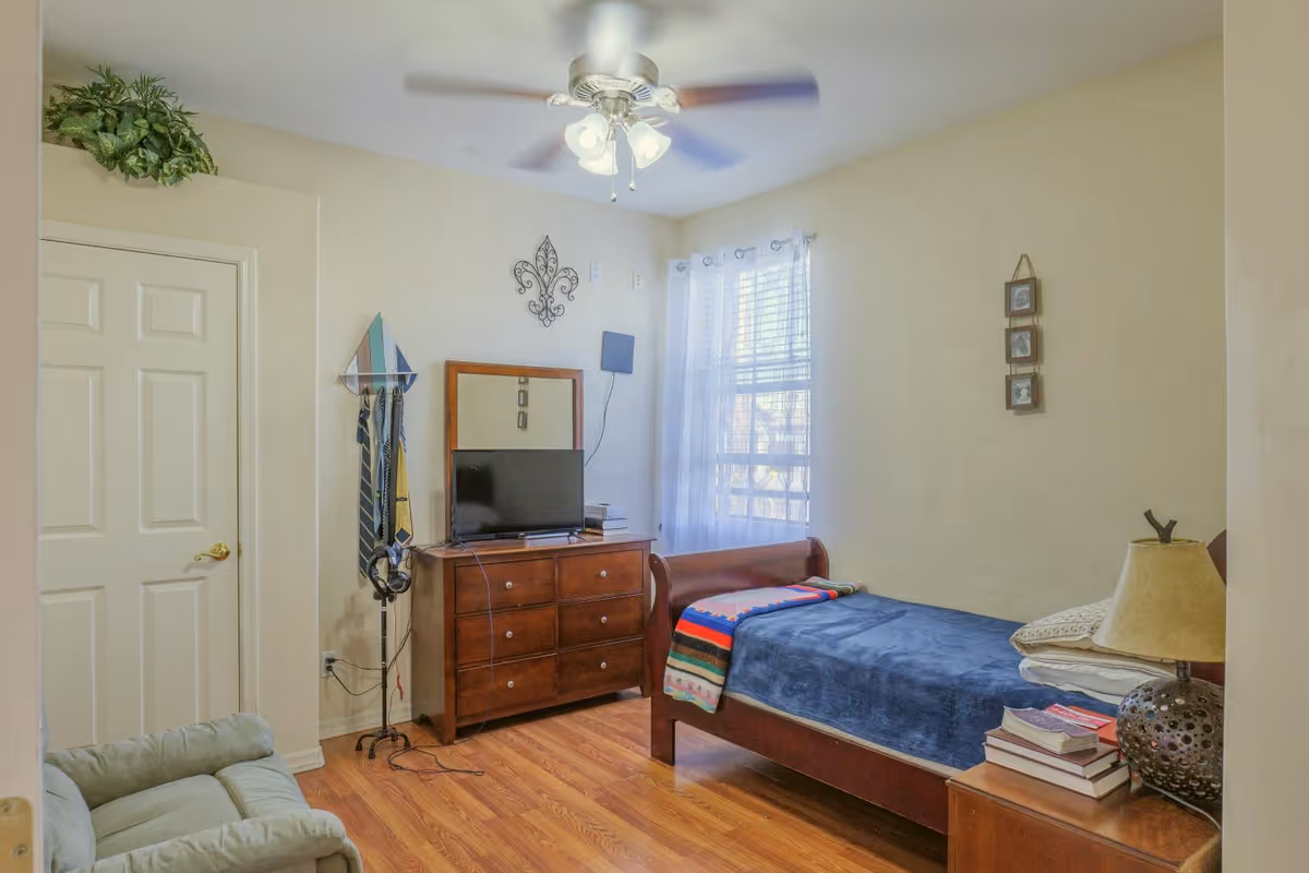 A tidy bedroom with a single wooden bed with a blue blanket, dresser with a TV, window with sheer curtains, ceiling fan, and a small armchair.