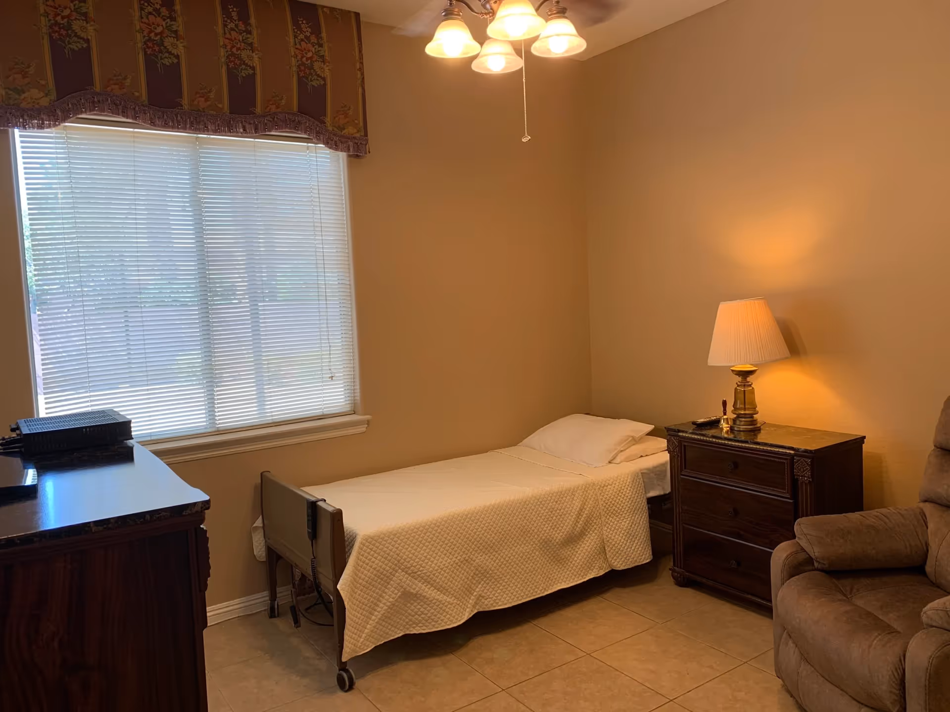 A furnished bedroom in a senior living facility with a bed, dresser, lamp, and recliner.