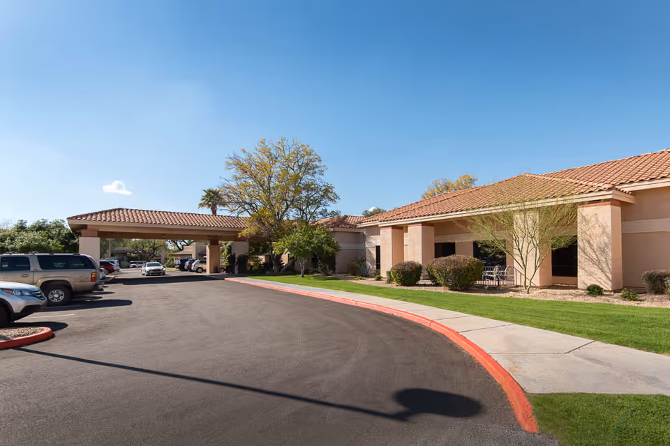 Front entrance and driveway of a single-story senior living center with a covered porte-cochere, parked cars, and landscaped lawn.