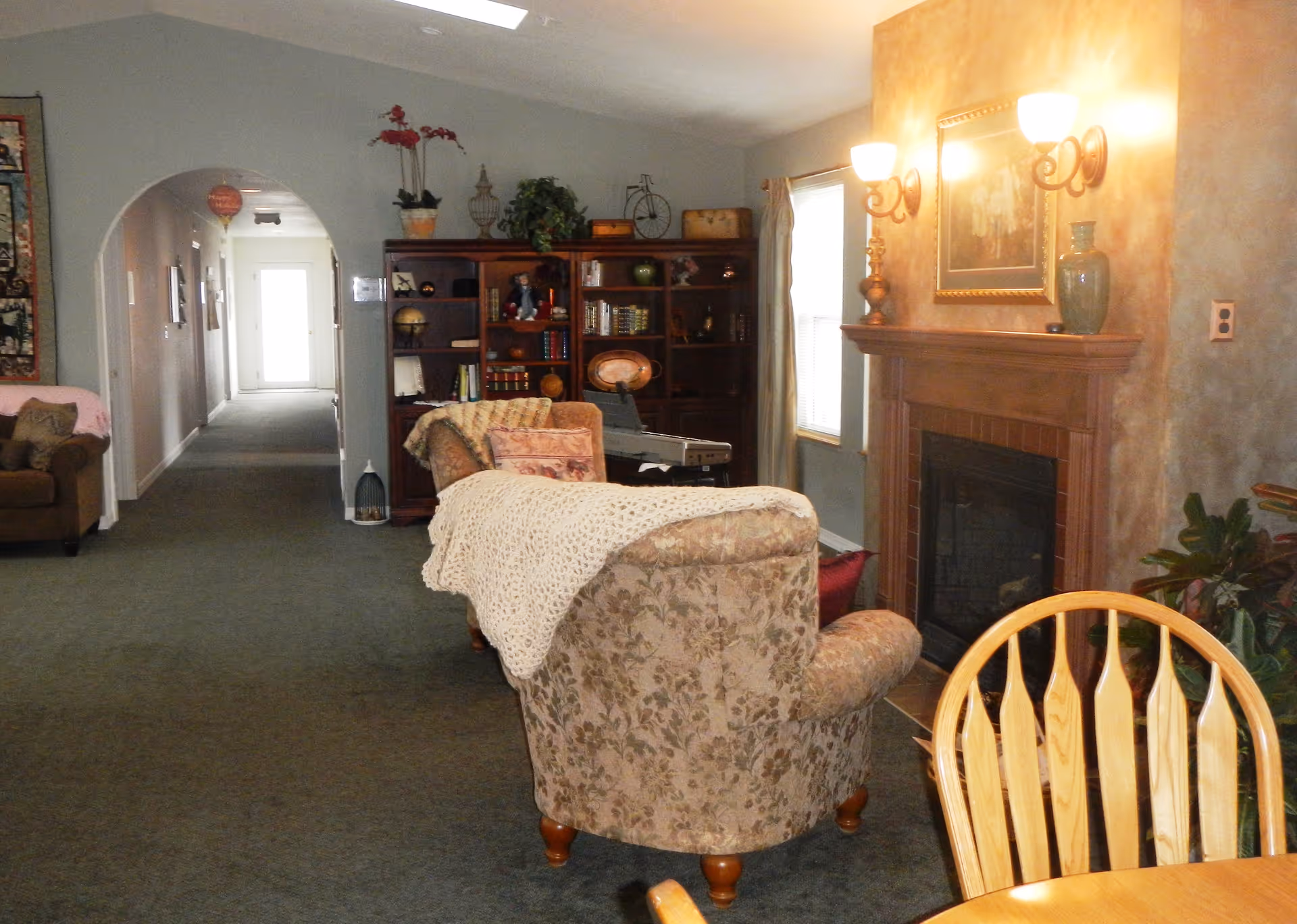 A cozy living room area in an assisted living facility with floral upholstered armchairs, a wooden bookshelf filled with books and decorative items, a fireplace with wall sconces above it, and a wooden dining table with chairs in the foreground. A hallway with an arched doorway leads to a bright door at the end.
