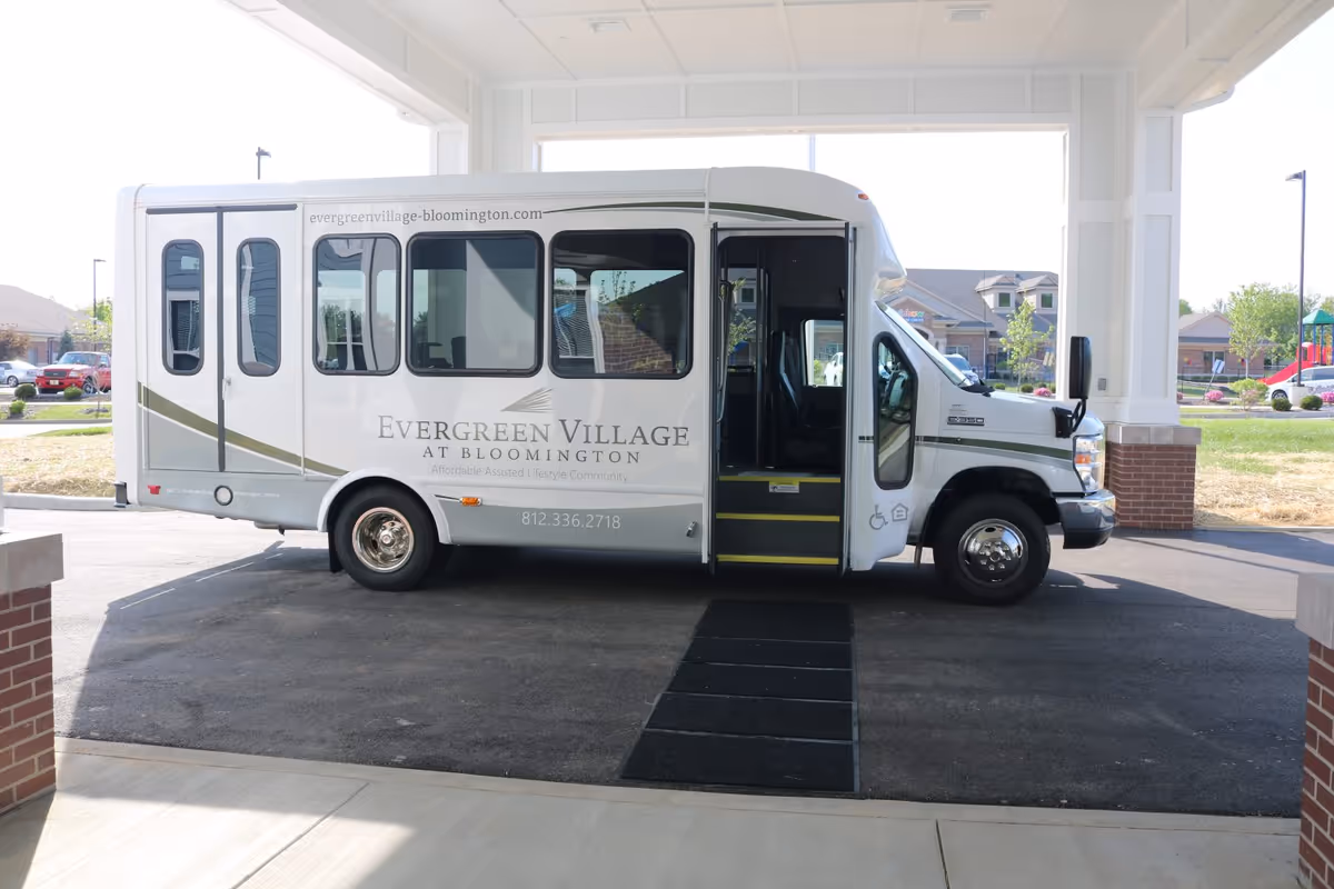 A white Evergreen Village shuttle bus parked under the entrance canopy of a senior living community.