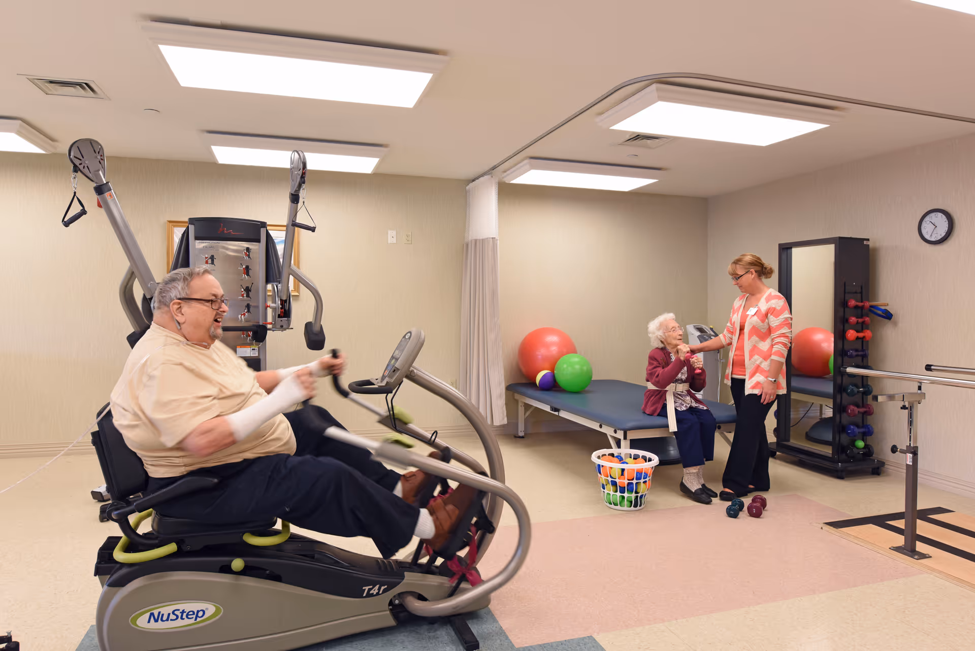 An elderly man exercises on a NuStep T4r machine in a physical therapy room while an elderly woman sits on a therapy table talking to a caregiver. The room contains exercise balls, dumbbells, a basket of balls, a large mirror, and parallel bars for walking exercises.