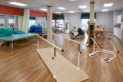 A spacious rehabilitation room with wooden flooring featuring parallel bars for walking exercises, exercise balls, therapy tables, an exercise bike, and various physical therapy equipment. The room has large windows with red and green valances, white walls, and ceiling lights providing bright illumination.