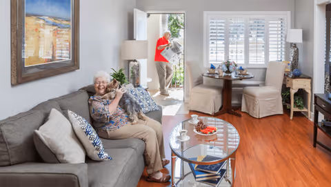 A cozy living room with a gray sofa where an elderly woman is sitting and smiling while holding a small dog. In the background, an elderly man is standing near an open door reading a newspaper. The room has wooden floors, a glass coffee table with books and a plate of grapes, a round dining table with two chairs, a lamp, and a large window with white shutters.