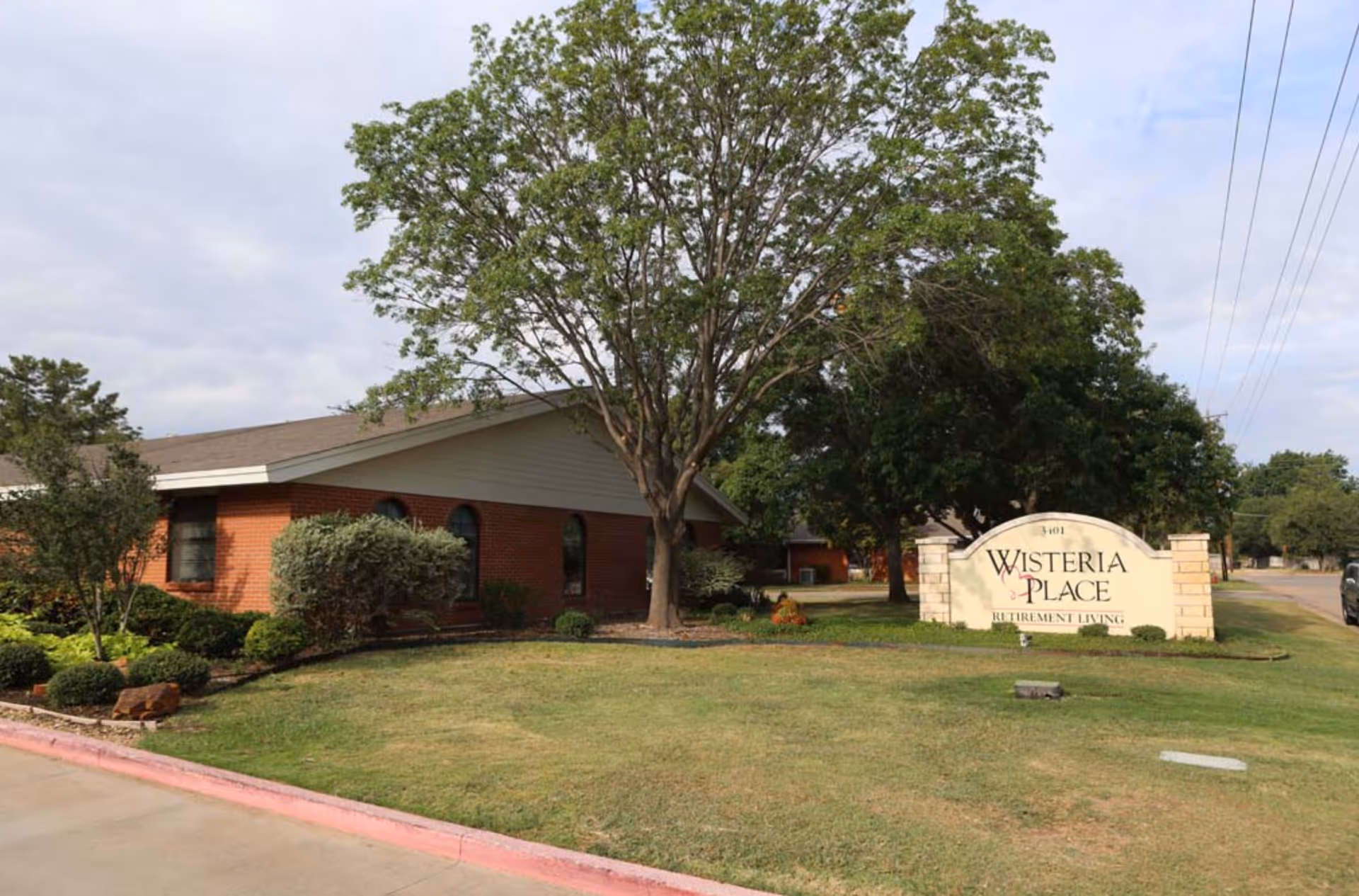Front exterior of a brick assisted living building with a large tree, lawn, and a sign reading "Wisteria Place".