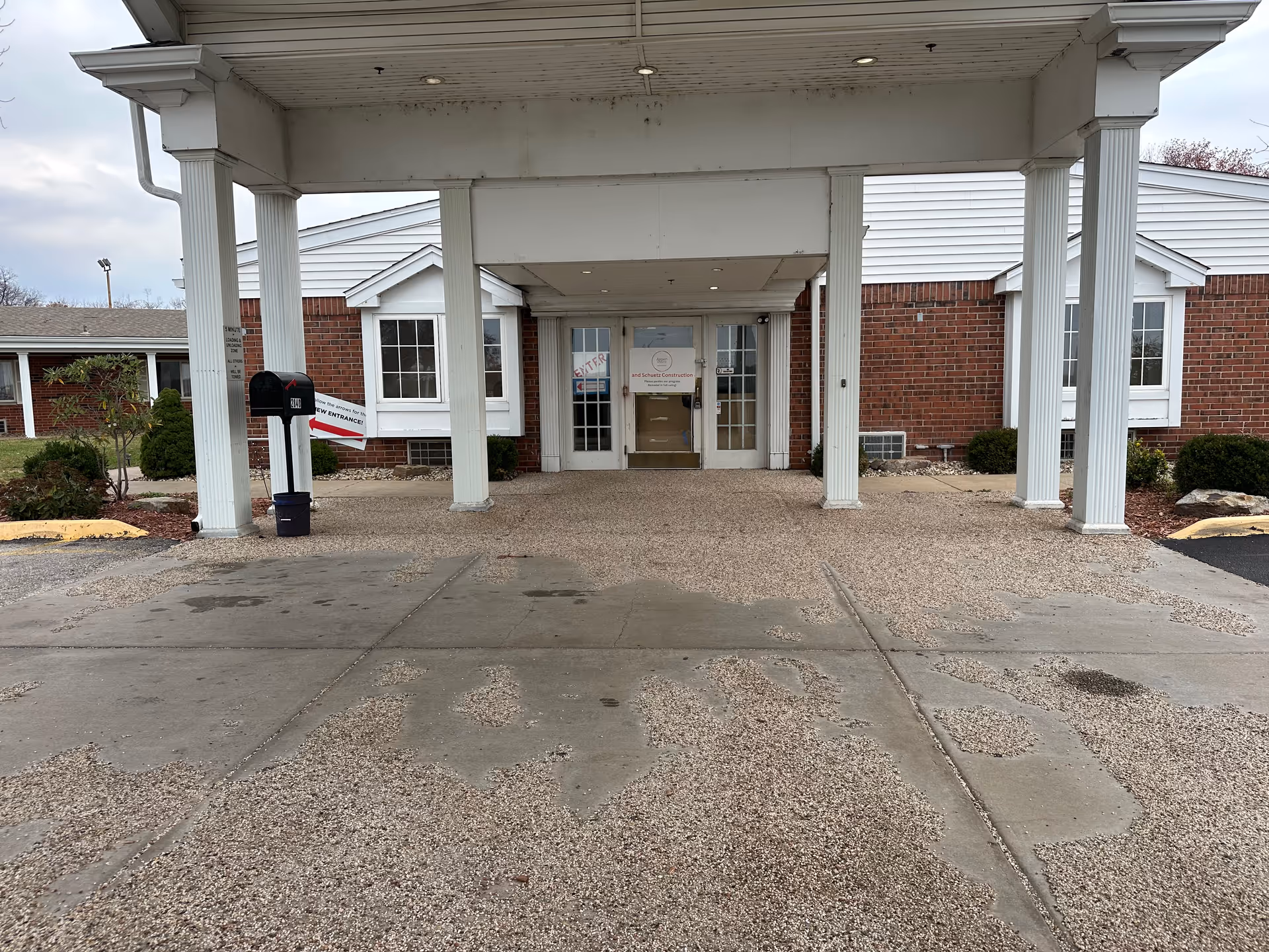 Covered entrance area of a brick building with white columns and a white ceiling. There are double glass doors at the entrance with signs on them, and a black mailbox on the left side near a sign pointing to a new entrance. The ground is a mix of concrete and textured pavement with some wet patches.