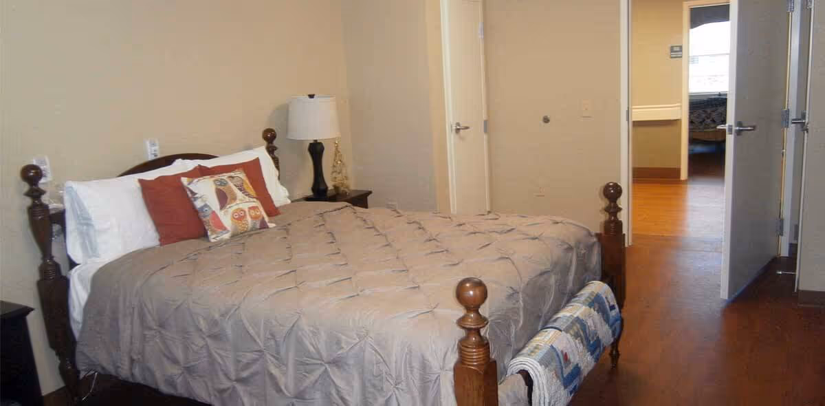 A furnished bedroom with a wooden four-poster bed, nightstand and lamp, and an open doorway into a hallway.