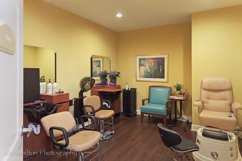 A small salon room with two beige salon chairs in front of mirrors and a hair washing station on the left. There is a teal armchair and a beige pedicure chair on the right side of the room. The walls are painted yellow, and there is a framed floral painting on the back wall. The floor is dark wood, and there are some plants and salon supplies on small tables and counters.