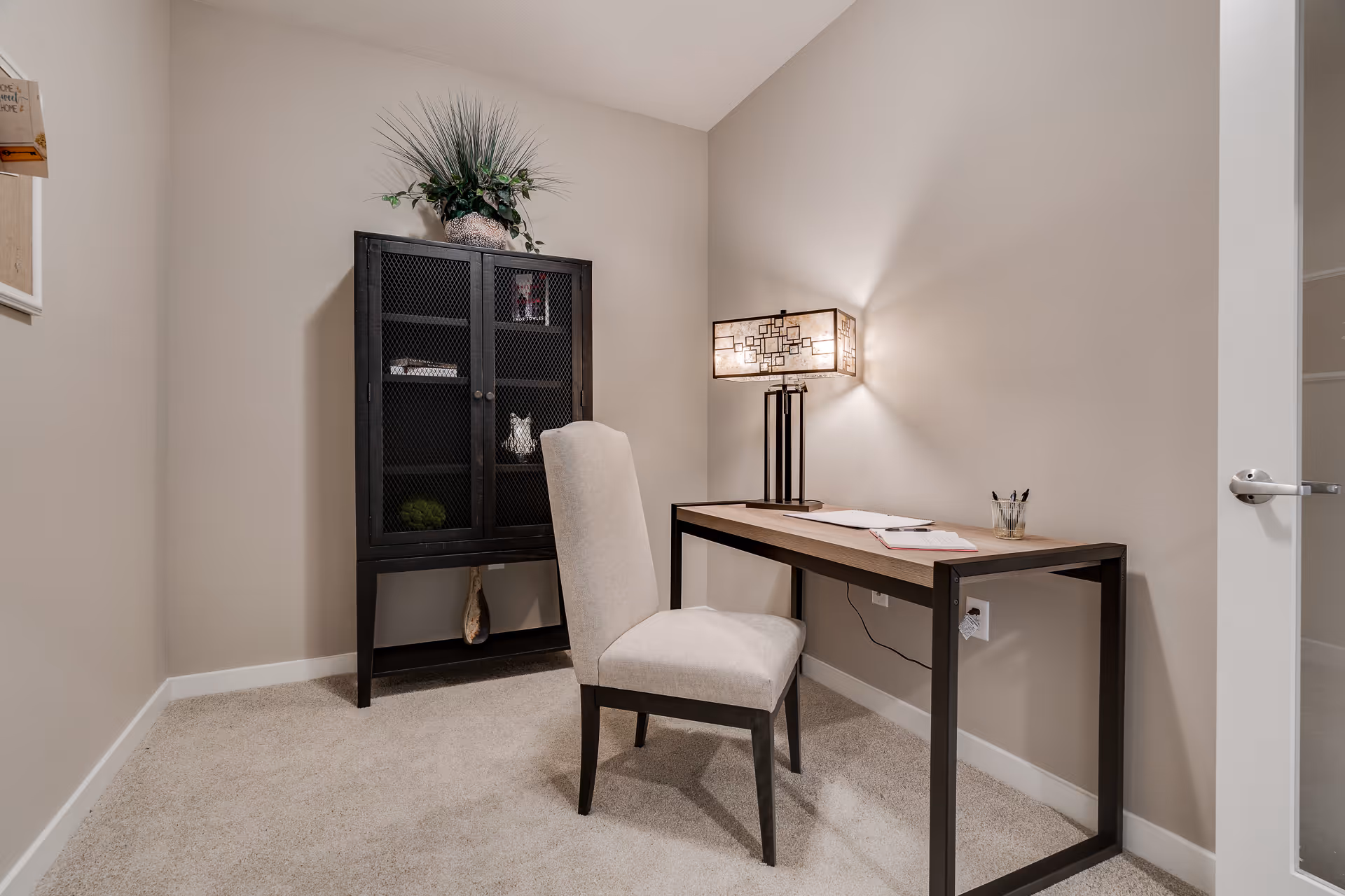 Small home office with a desk, upholstered chair, black storage cabinet, and lamp against beige walls.