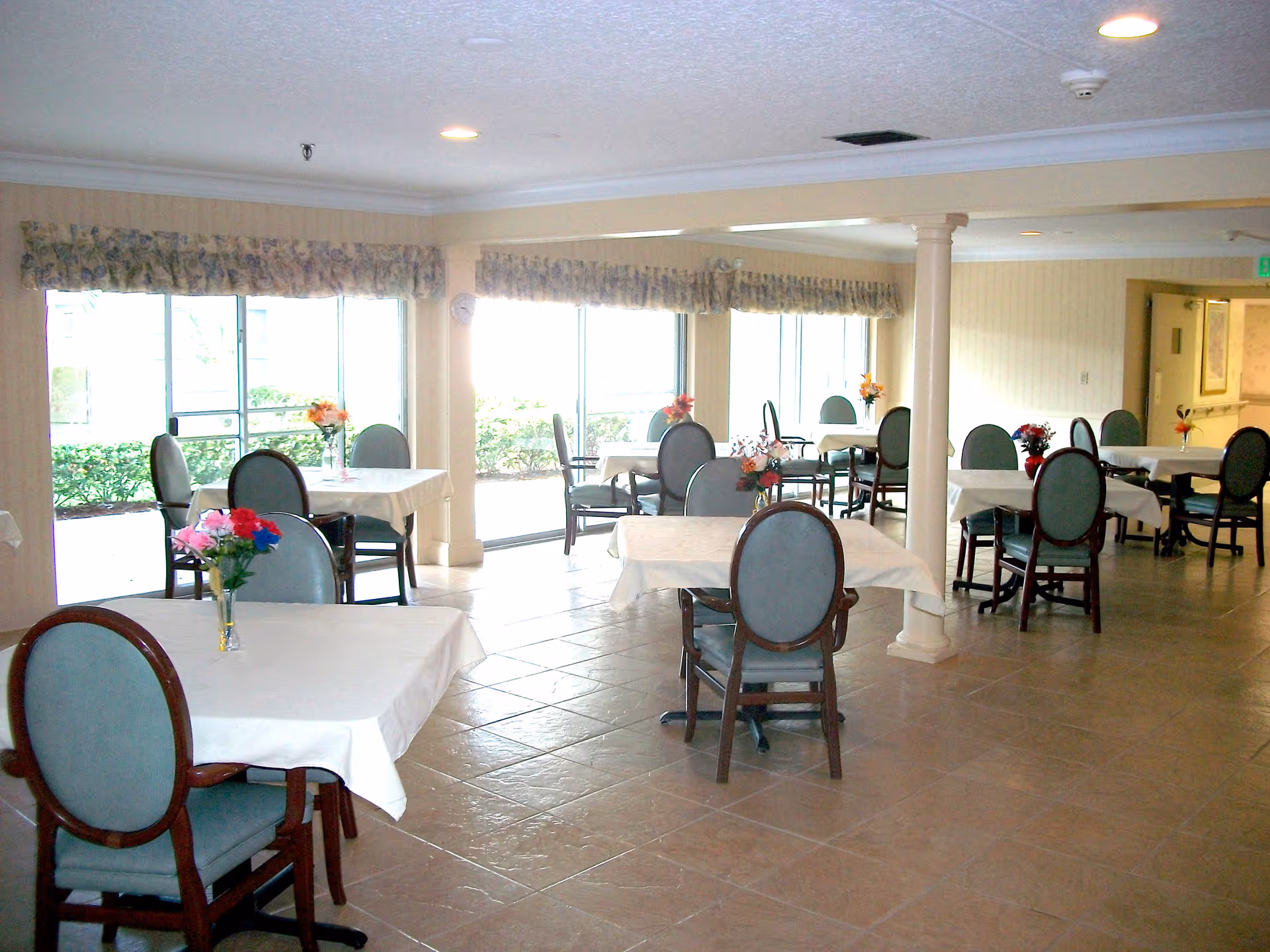 A bright dining room with multiple tables covered in white tablecloths, each adorned with small flower arrangements. The room has large windows with floral valances, letting in natural light. Chairs with blue upholstery and wooden frames are arranged around the tables. The floor is tiled, and there are columns supporting the ceiling.
