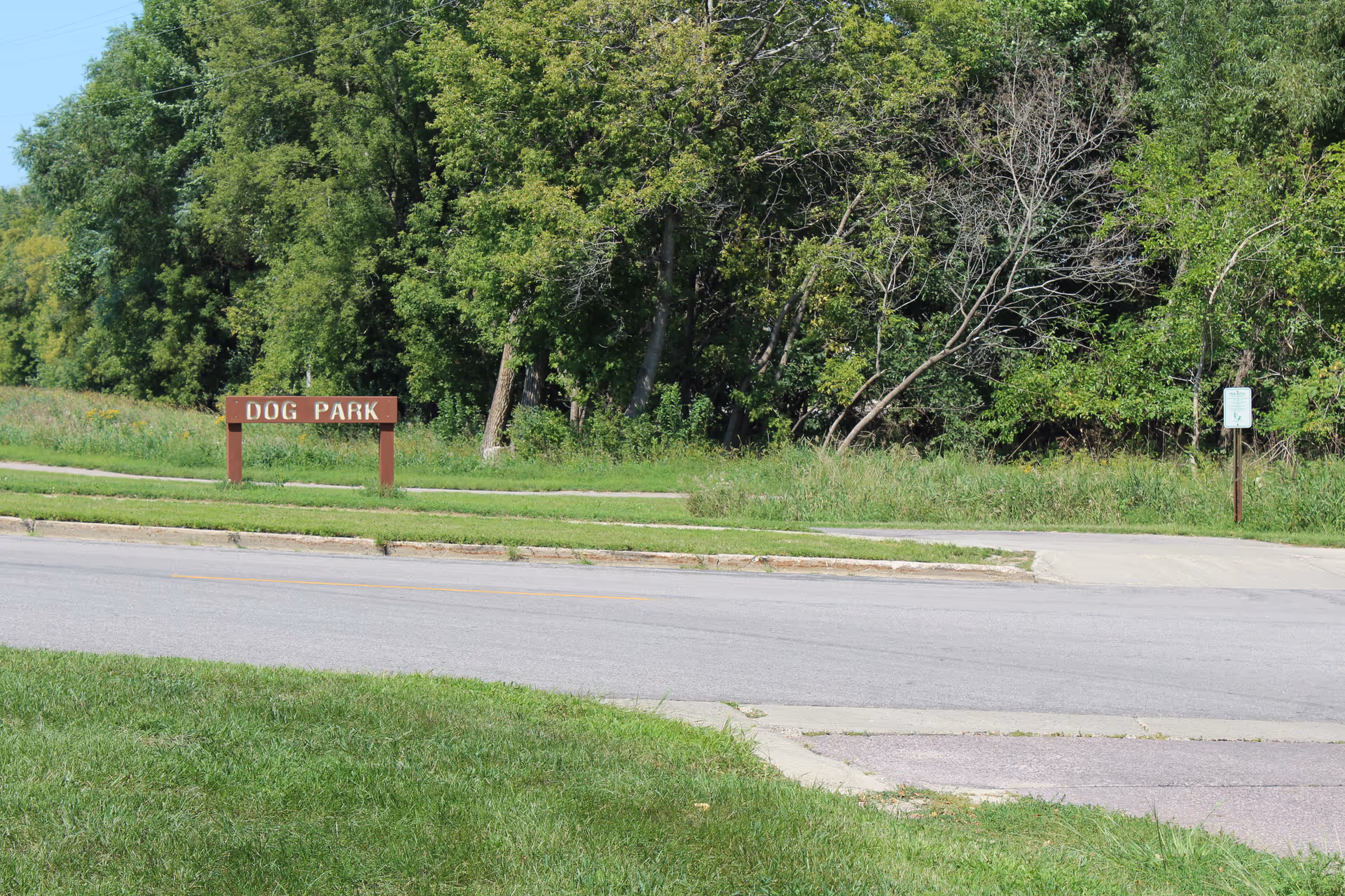 Grassy roadside area with a wooden 'DOG PARK' sign and dense trees in the background.