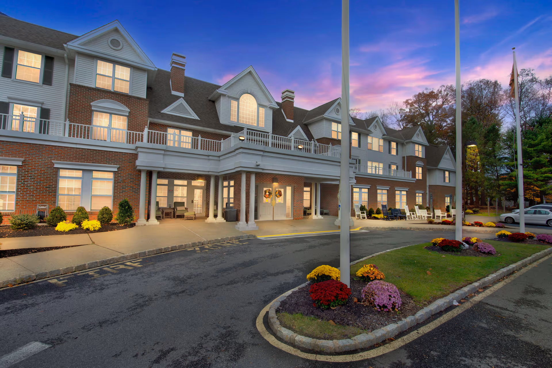 Exterior view of Brighton Gardens of Florham Park senior living facility at dusk, showing a large brick and white-paneled building with multiple windows, a covered entrance with columns, landscaped flower beds with colorful flowers, and a driveway with flagpoles.