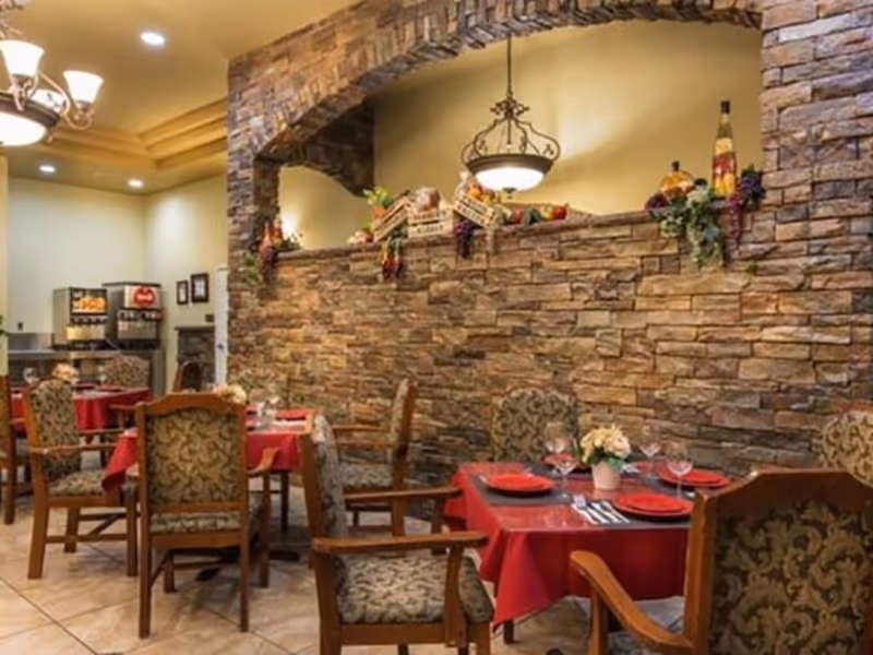 Dining area with tables covered in red tablecloths, set with plates, glasses, and silverware. The room features a stone wall with an arched opening decorated with artificial fruits and greenery. Upholstered chairs with wooden frames surround the tables. In the background, there is a beverage station with dispensers.