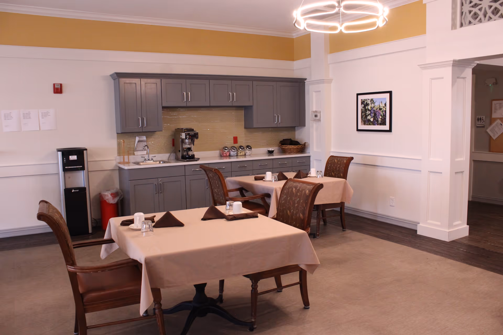 A dining area with two tables covered in beige tablecloths, each set with brown napkins, cups, and glasses. There are four wooden chairs with patterned cushions around the tables. In the background, there is a kitchenette with gray cabinets, a coffee machine, and a water dispenser. The walls are white with a yellow accent near the ceiling, and a framed floral picture hangs on the wall. A modern circular light fixture hangs from the ceiling.