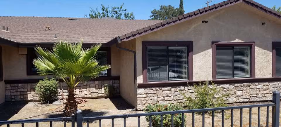Front exterior of a single-story building with a stone lower facade, three windows with blinds, a small palm tree and a metal fence.