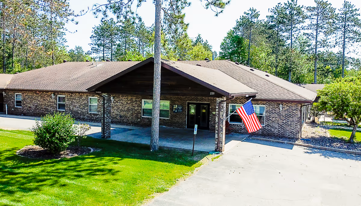 Exterior view of a single-story brick building with a covered entrance, an American flag mounted near the entrance, surrounded by green grass, trees, and a clear blue sky.