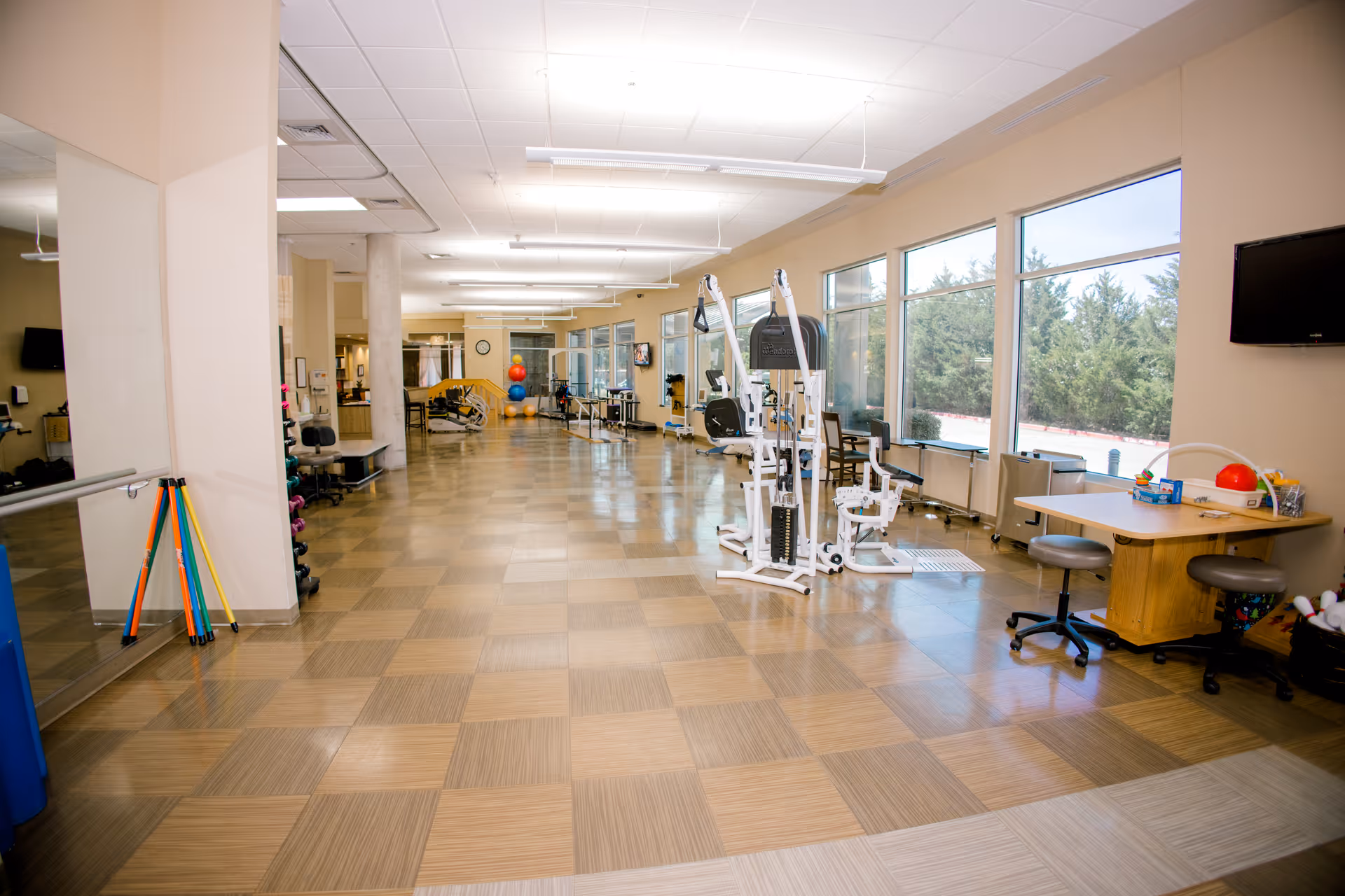 A spacious rehabilitation and wellness center room with large windows along one side letting in natural light. The room features exercise equipment, therapy balls, a desk with chairs, and various therapy tools. The floor has a checkered pattern, and the walls are painted a light beige color.