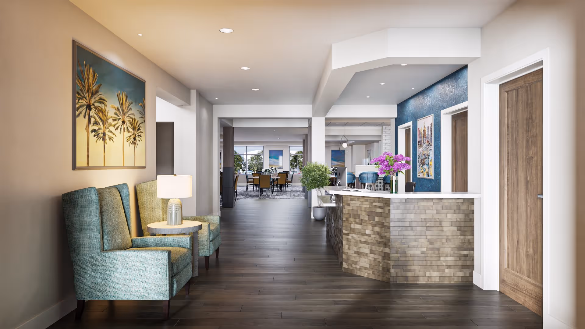 Interior view of a senior living facility hallway with two teal armchairs and a small round table with a lamp on the left. A reception desk with a brick facade and a vase of purple flowers is on the right. The hallway leads to a dining area with tables and chairs, and there are framed artworks on the walls.