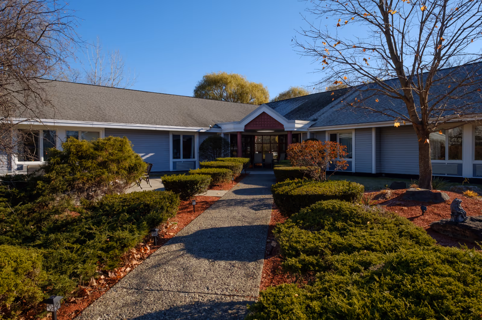 Exterior view of a single-story building with a gray roof and blue siding, surrounded by neatly trimmed bushes and trees with some autumn leaves. A paved walkway leads to the entrance, which has a small covered porch with red accents.