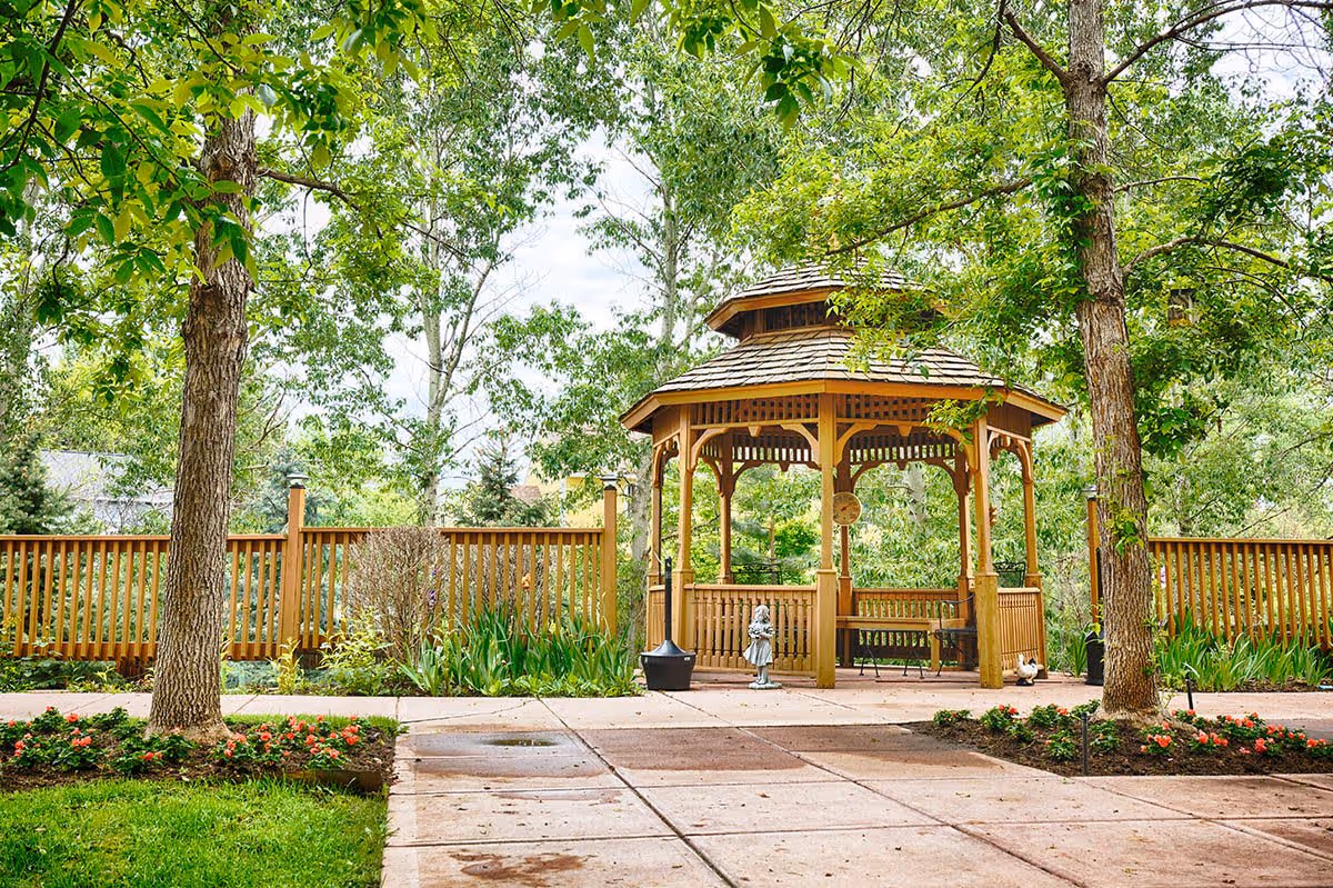 A wooden gazebo surrounded by trees and greenery in a garden area with a paved walkway and flower beds, located within a fenced outdoor space.