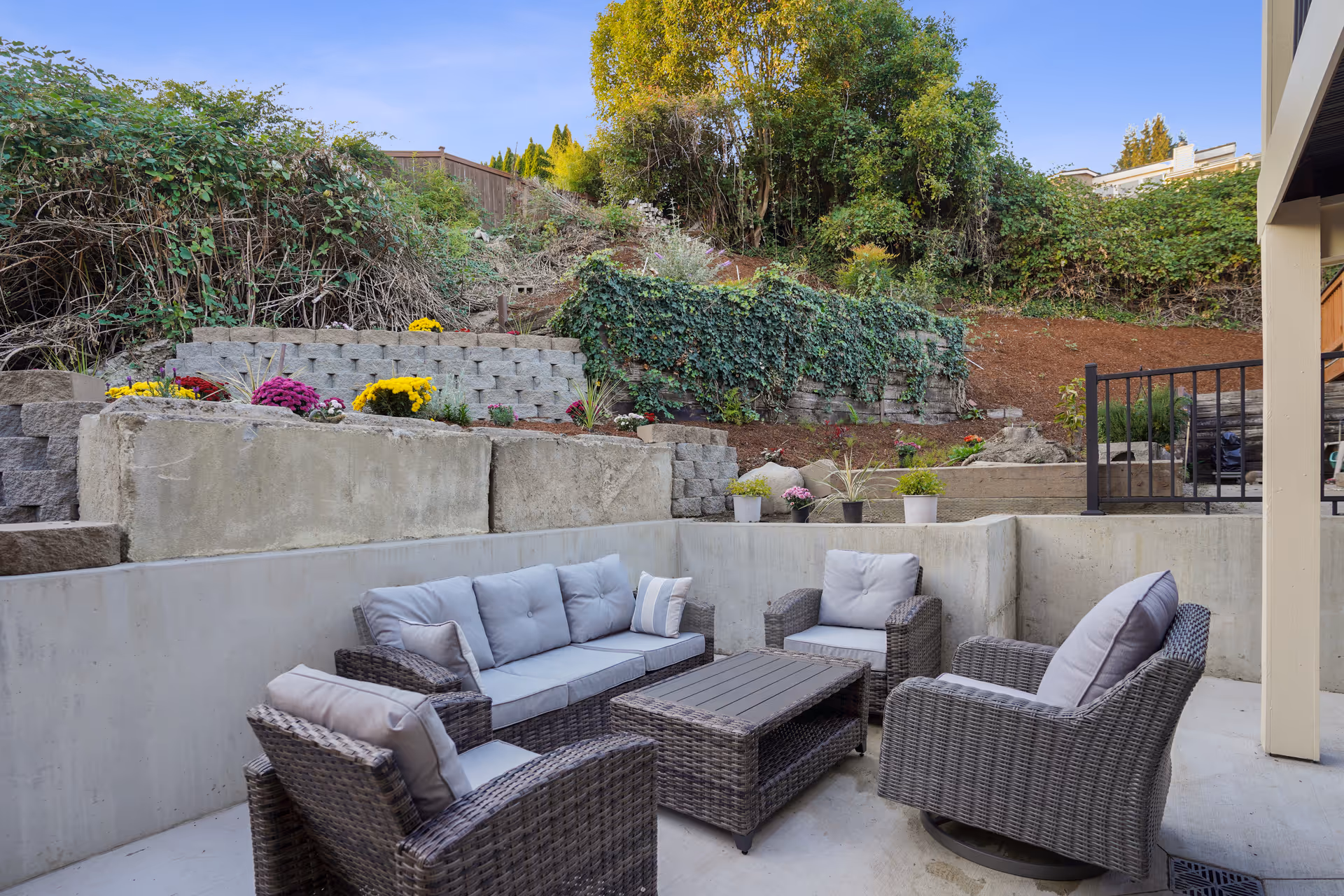 Outdoor patio area with a set of gray cushioned wicker furniture including a sofa, three armchairs, and a rectangular coffee table. The patio is surrounded by concrete walls and a landscaped hillside with various plants and flowers. Trees and bushes are visible in the background under a clear blue sky.
