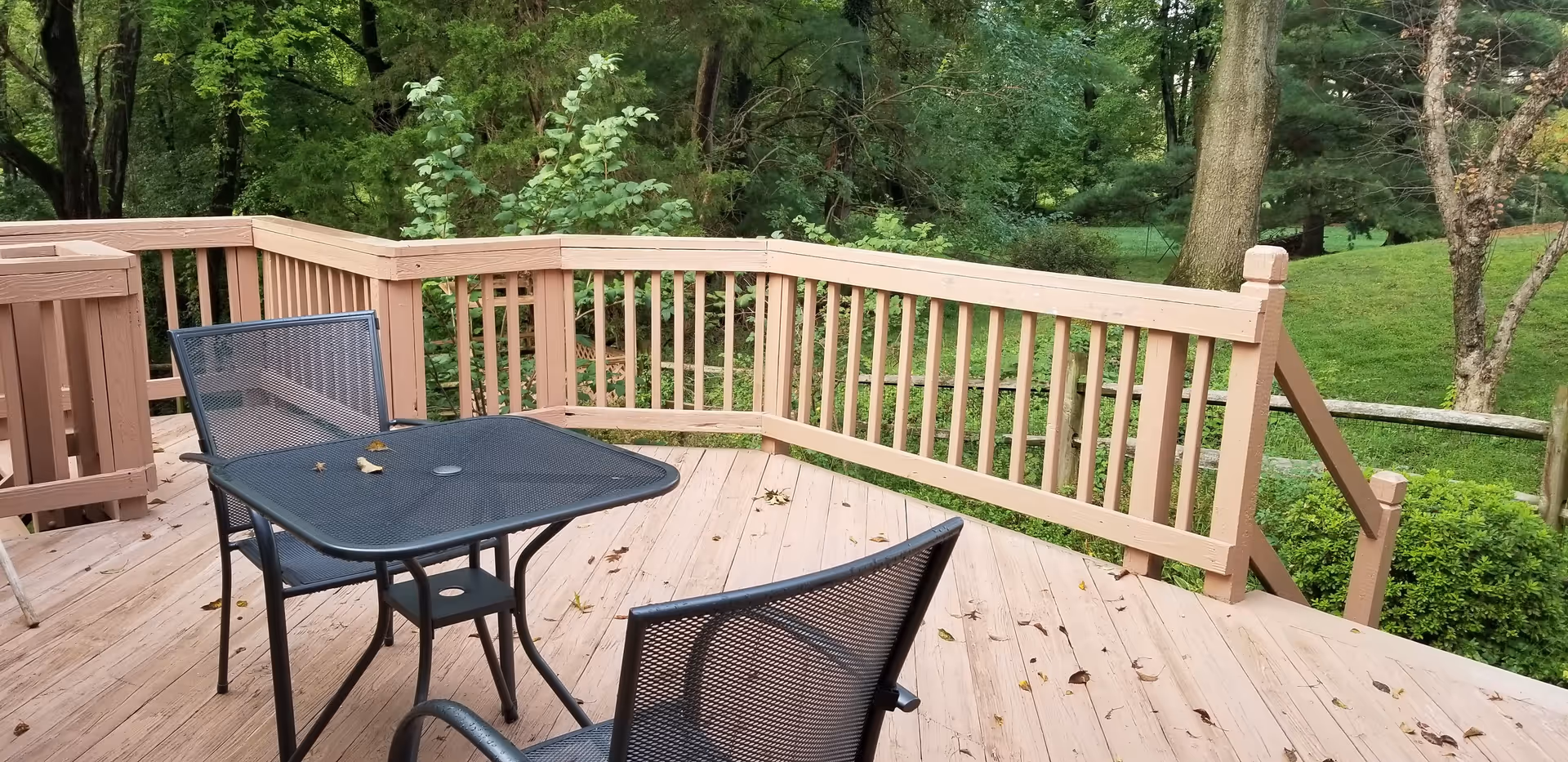 Outdoor wooden deck with a black metal table and two matching chairs, surrounded by a wooden railing. The deck overlooks a green, wooded area with trees and bushes.