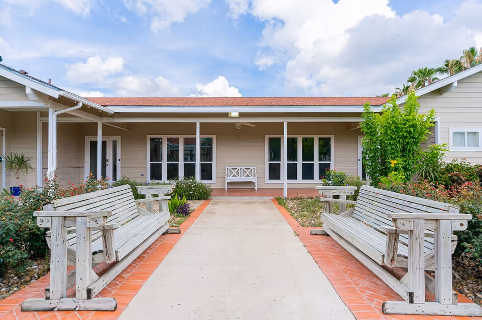 Front entrance of a single-story assisted living building with a paved walkway flanked by wooden benches and garden beds.