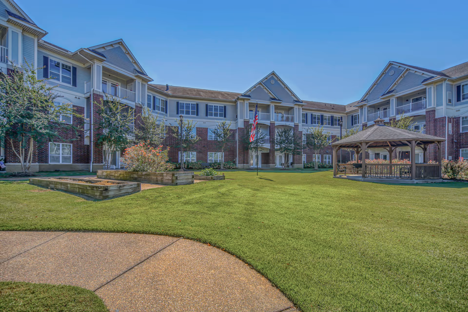 Outdoor courtyard area of Irene Woods Assisted Living featuring a well-maintained lawn, a wooden gazebo with benches, raised garden beds, trees, and a three-story building with balconies and windows surrounding the courtyard under a clear blue sky.