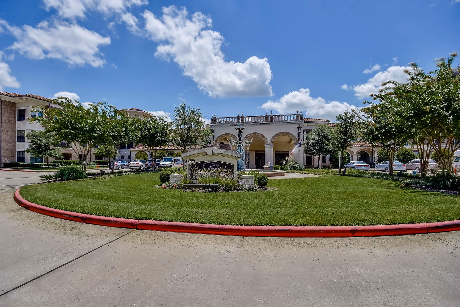 Front exterior view of Conservatory At Champion Forest senior living facility with a circular driveway, green lawn, trees, and a fountain under a partly cloudy blue sky.