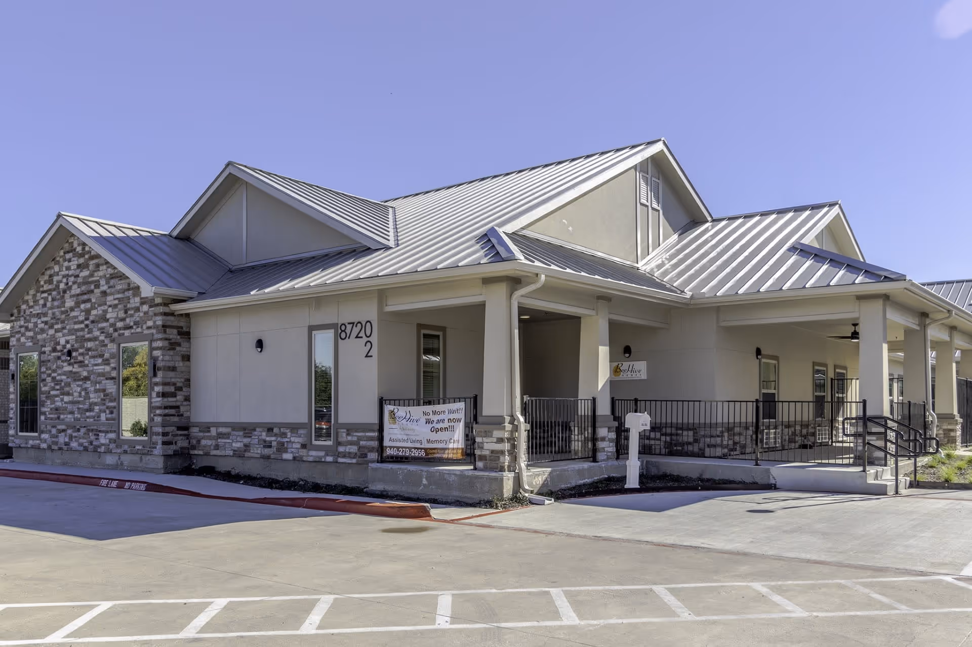 Exterior view of a single-story building with a metal roof and stone accents on the walls. The building has a covered porch with black railings and a ramp leading to the entrance. There is a sign on the porch that reads 'BeeHive Homes of McKinney' and another banner indicating the facility is now open for assisted living and memory care. The parking area in front has marked spaces and a fire lane.