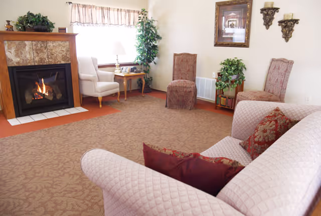 A cozy living room with a lit fireplace on the left, a white armchair next to a wooden side table with a lamp, two upholstered chairs against the wall, a framed picture, decorative wall sconces, and a patterned sofa with red and gold cushions in the foreground.