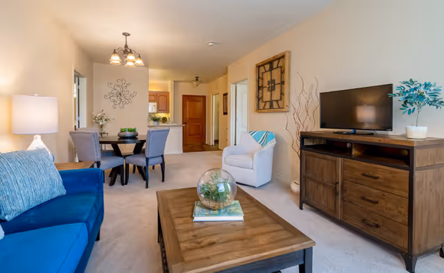 A cozy living room and dining area in a senior living facility with a blue sofa, a wooden coffee table with a glass terrarium and books, a white armchair with a blue throw, a wooden TV stand with a flat-screen TV and a potted plant, and a round dining table with four chairs. The walls are beige, and there is a decorative wall clock and wall art. The kitchen and hallway are visible in the background.