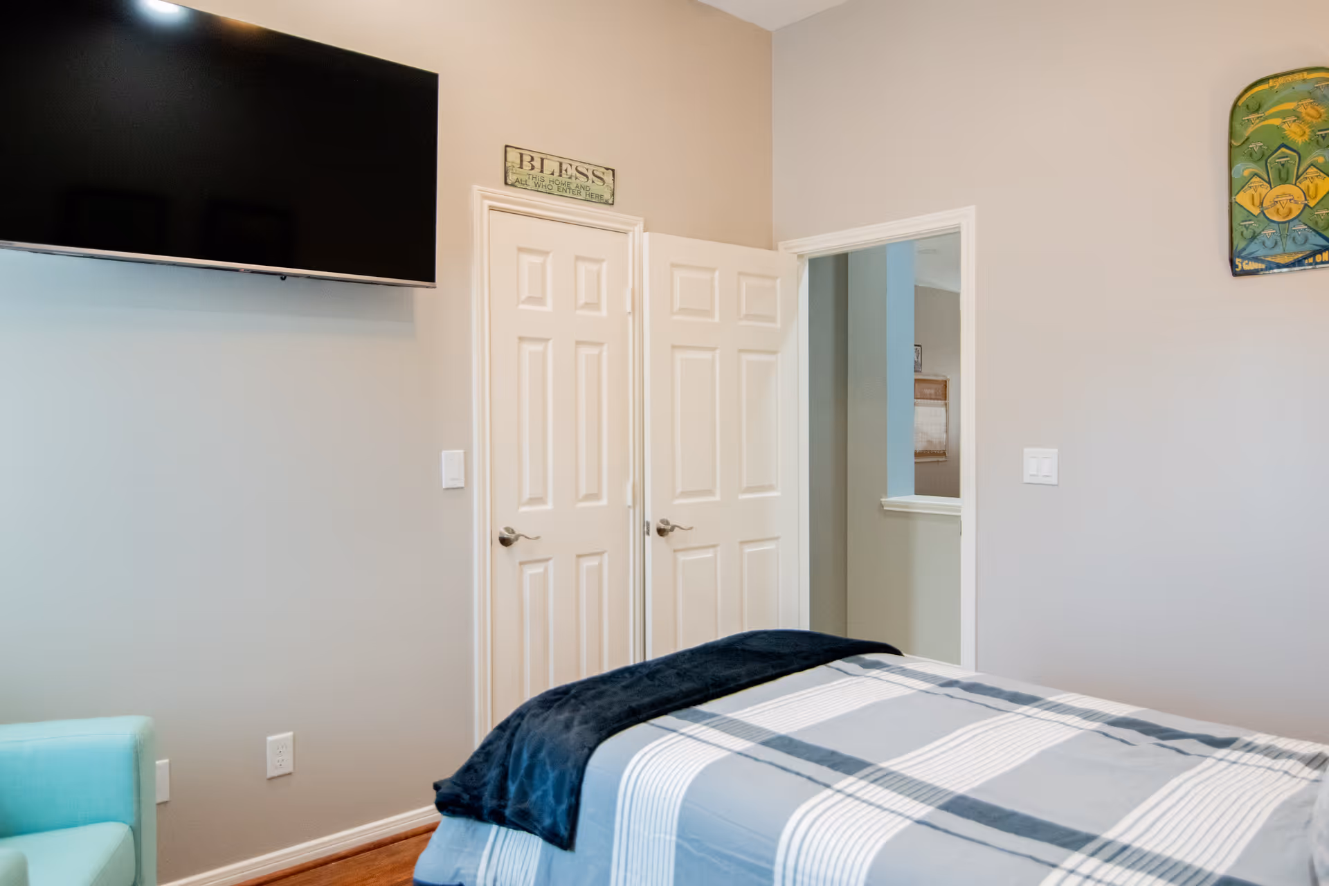 A bedroom with a bed covered in a gray and white striped blanket and a dark blue throw. There is a light blue armchair in the corner, a large flat-screen TV mounted on the wall, and two white doors, one slightly open. Above one door is a sign that reads 'BLESS THIS HOME AND ALL WHO ENTER HERE'. The walls are painted light beige.