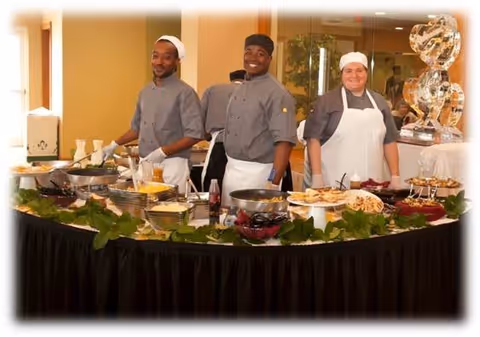 Three chefs standing behind a buffet table filled with various dishes and garnished with green leaves. The chefs are smiling and wearing gray chef jackets and white aprons and hats. The background shows a warm indoor setting with a large ice sculpture on the right side.