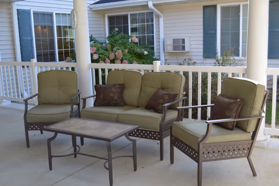 Covered outdoor patio with green cushioned chairs, a loveseat, and a coffee table in front of a white railing and building facade.