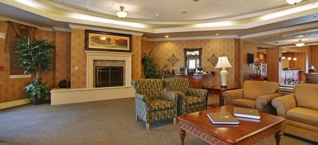 A cozy living room area with patterned armchairs, a sofa, a wooden coffee table with books on it, a fireplace with a framed painting above it, a large potted plant, and warm lighting from a table lamp and ceiling fixtures. The room has beige walls with a subtle pattern and carpeted flooring. In the background, there is a view into a kitchen area with bar stools and pendant lights.