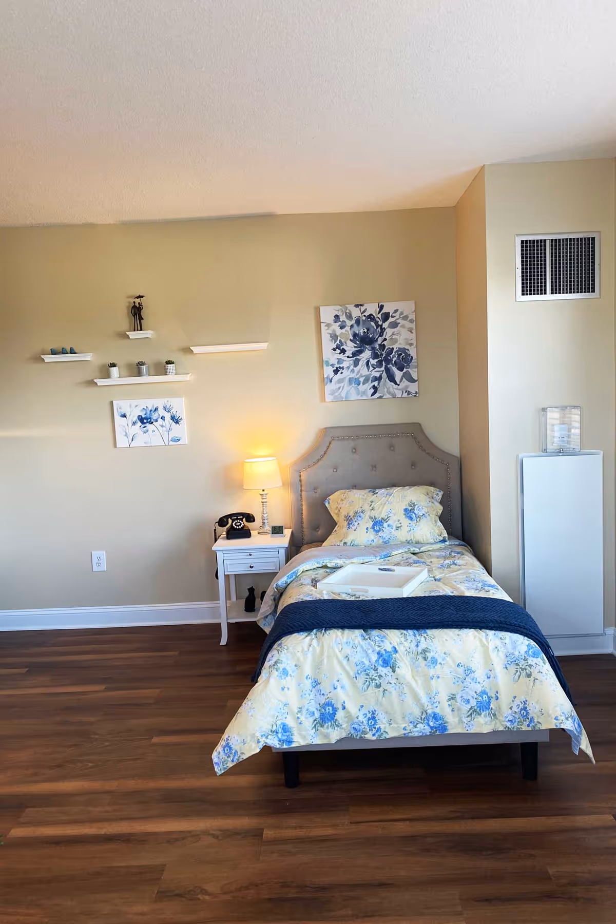 A single bed with a floral blue and yellow bedspread and matching pillow is placed against a beige wall. Above the bed are two floral paintings in blue tones. To the left of the bed is a white nightstand with a vintage black rotary phone, a small lamp with a beige shade, and a small clock. The floor is wooden, and there are small white shelves on the wall with decorative items including small plants and figurines.