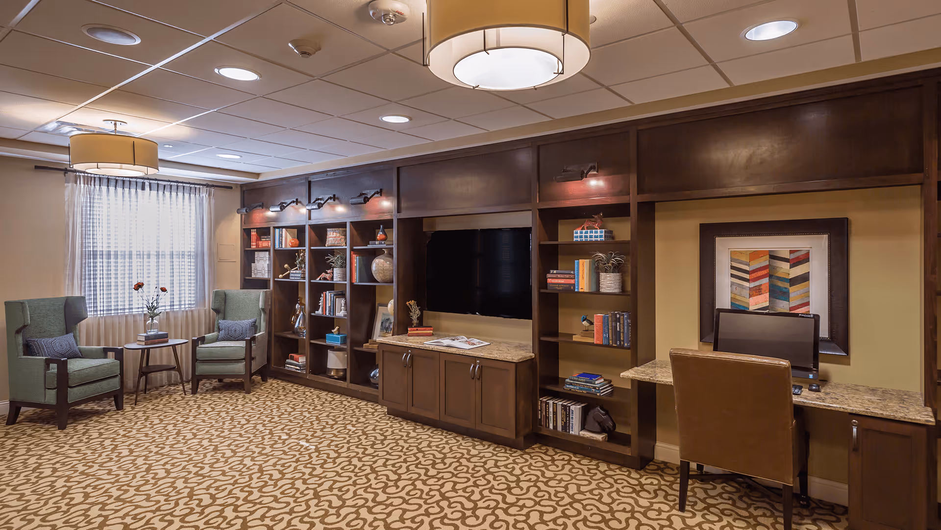 A cozy common area with two green armchairs and a small round table with flowers near a window with sheer curtains. The wall features built-in dark wood shelves filled with books and decorative items, a flat-screen TV, and a small desk with a computer and a brown leather chair. The room has patterned carpet and ceiling lights with round fixtures.