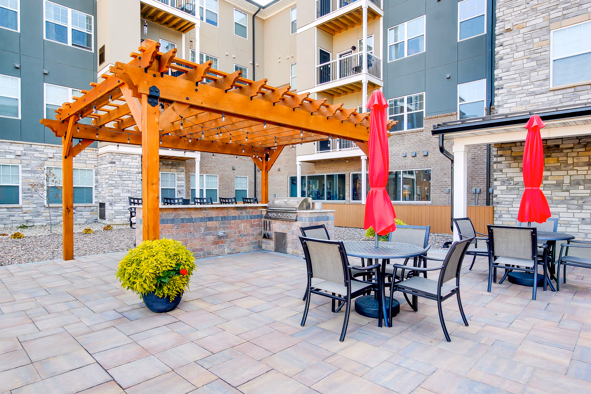 Outdoor courtyard patio with a wooden pergola, round dining tables with red umbrellas, a potted plant, and the apartment building facade in the background.