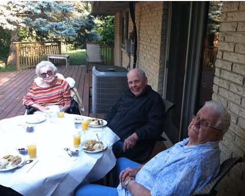 Three elderly people sitting around a round table outdoors on a wooden deck, enjoying a meal with plates of food and glasses of orange juice. The setting is next to a brick building with a sliding glass door and an air conditioning unit visible. Trees and greenery are in the background.