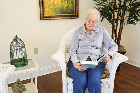 An elderly woman sits in a white wicker chair reading a book in a bright sitting area with a framed painting, potted plant, and side table.