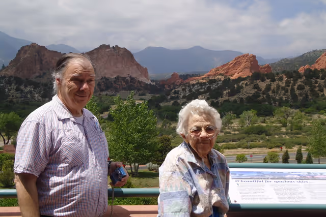 An elderly man and woman standing outdoors with a scenic view of red rock formations, green trees, and mountains in the background under a partly cloudy sky. The man is holding a camera and both are smiling at the camera.