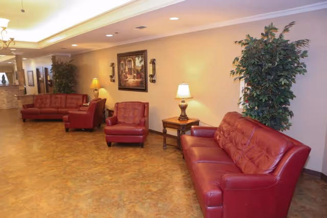 A well-lit interior lounge area with red leather sofas and armchairs arranged along the walls. There are two table lamps on wooden side tables, a large framed painting on the wall, and a tall green potted plant in the corner. The floor has a warm-toned pattern, and the ceiling features recessed lighting and a chandelier.