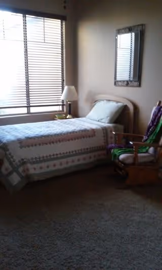 Small bedroom with a single bed beside a large window with blinds, a bedside lamp, wall mirror, and a wooden rocking chair.