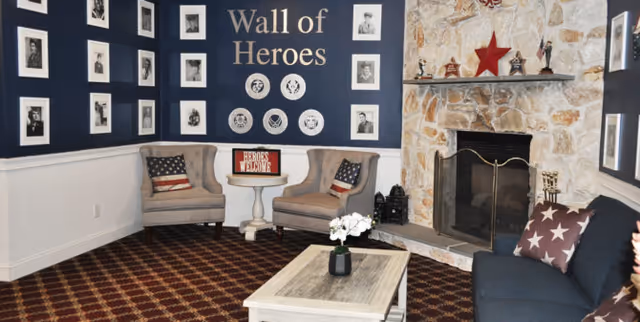 A cozy living room area featuring a stone fireplace with a red star decoration on the mantle. Two beige armchairs with American flag pillows flank a small round table with a 'Heroes Welcome' sign. A dark blue wall labeled 'Wall of Heroes' displays framed black and white photos and circular portraits. A dark blue sofa with star-patterned pillows and a light wooden coffee table with a small plant complete the seating area. The floor is covered with a patterned carpet.