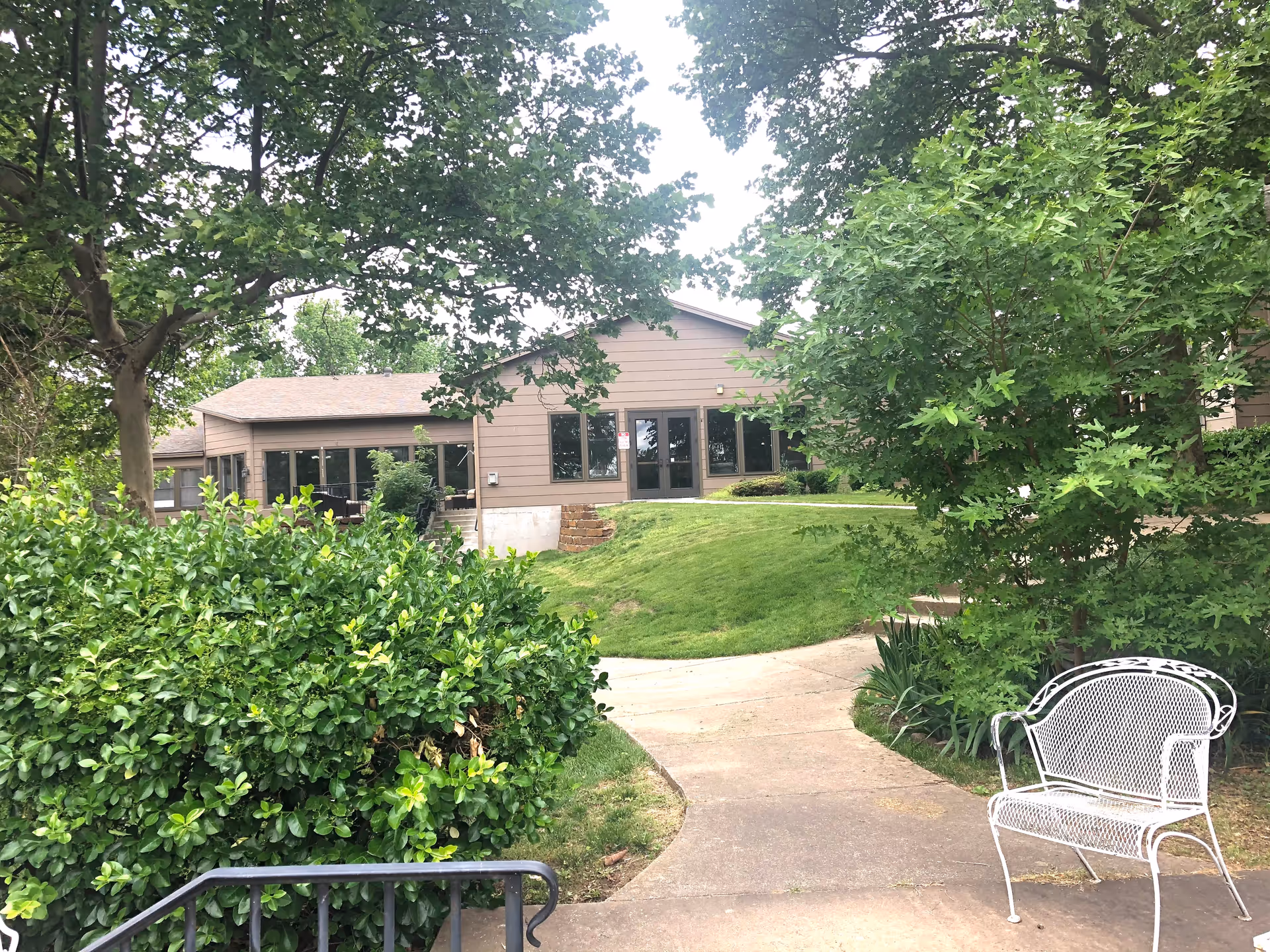 Outdoor view of a retirement community building surrounded by lush green trees and bushes. A paved walkway leads up to the building entrance, and a white metal bench is placed on the right side near the path.