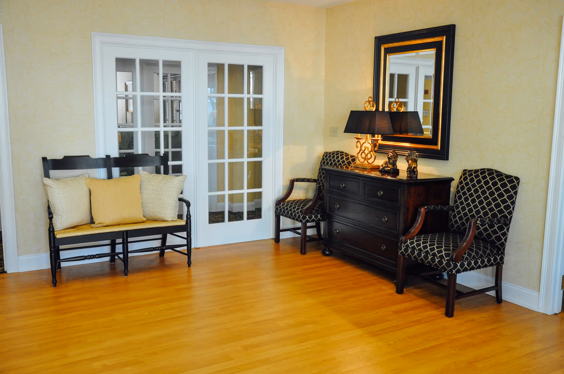 A cozy interior room with wooden flooring, featuring a black wooden bench with three cushions on the left, a dark wooden dresser with a lamp and decorative items on top, a large framed mirror above the dresser, and two patterned armchairs on either side of the dresser. The background includes white-framed glass double doors.